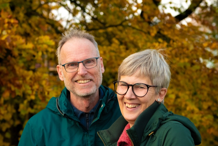 Två personer i gröna jackor framför ett träd med höstlöv. Foto.